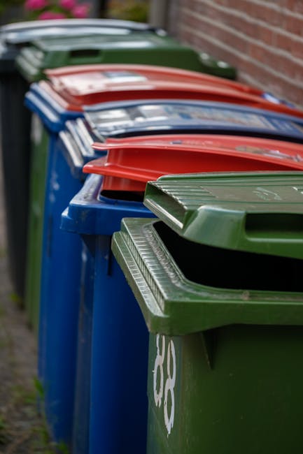 A black wheeled rubbish bin labeled with St. John's is positioned on a sidewalk beside a street at night. The lid of the bin is open, revealing assorted waste items including cardboard boxes and plast