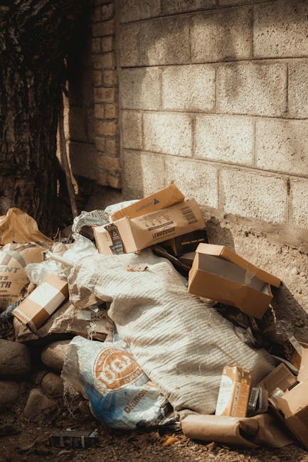 Two large black plastic bags filled with rubbish are placed on the edge of a paved sidewalk, positioned against a black metal fence with vertical bars. The bags appear to be tightly knotted at the top